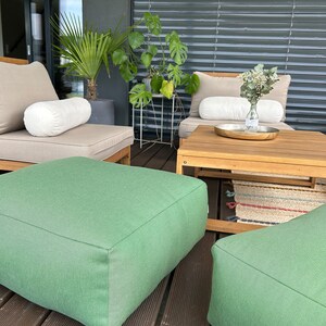 May include: Two green square ottomans on a wooden deck with a wooden table and two beige chairs with white cushions. The chairs are facing each other and there are plants in the background.