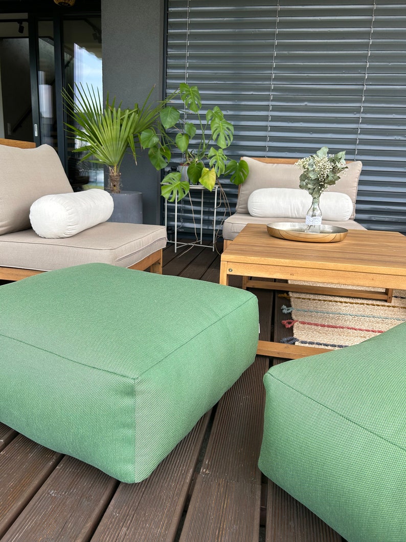 May include: Two green square ottomans on a wooden deck with a wooden table and two beige chairs with white cushions. The chairs are facing each other and there are plants in the background.