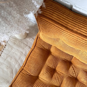 Op de afbeelding: Een mosterdgele corduroy vloercushion met een gestoffeerd ontwerp. Het kussen is gemaakt van een zachte, pluche stof en is perfect om elke kamer een vleugje comfort en stijl te geven.