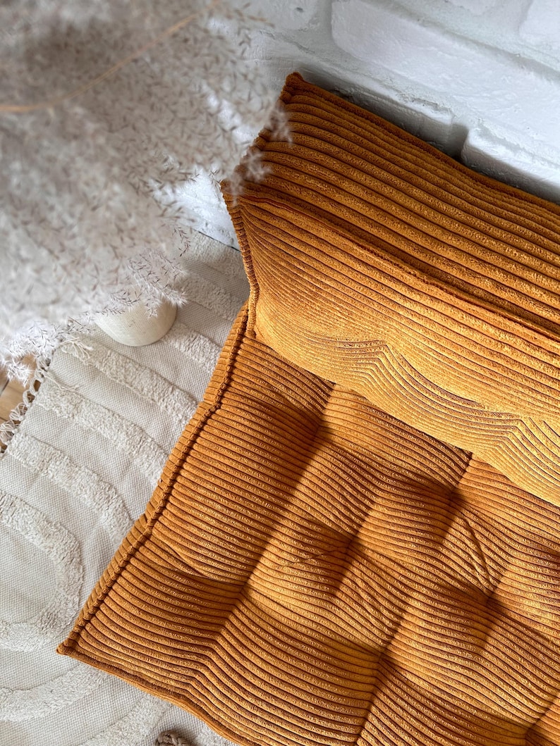 Op de afbeelding: Een mosterdgele corduroy vloercushion met een gestoffeerd ontwerp. Het kussen is gemaakt van een zachte, pluche stof en is perfect om elke kamer een vleugje comfort en stijl te geven.