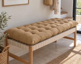 May include: A rectangular, tufted bench cushion in a solid tan color, featuring a thick, soft-filled construction with multiple button-tufted indentations across the surface, resting on a wooden bench frame with light-colored woven seat straps.