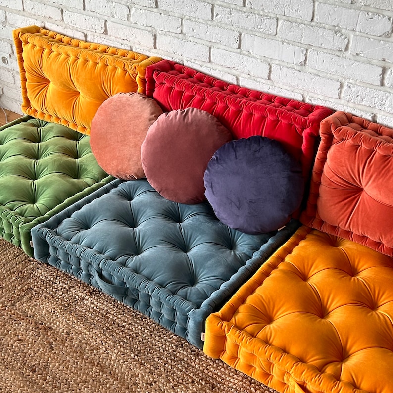 May include: A set of six floor cushions in various colors and textures. The cushions are made of velvet and have a tufted design. The cushions are arranged in a row on a woven rug.