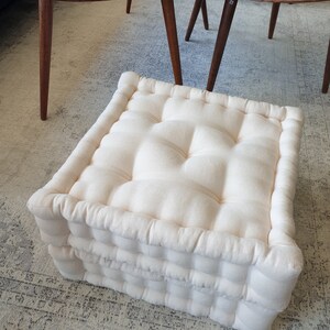 May include: A white, square, tufted floor cushion with a thick, padded design. The cushion is made of a soft, natural fabric and has a simple, elegant look.
