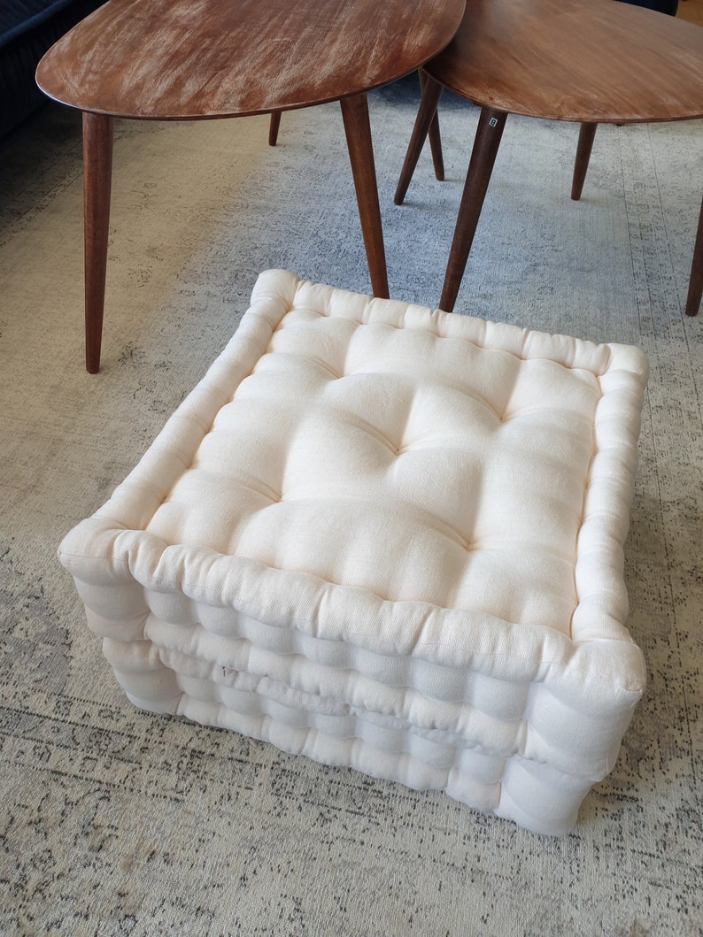 May include: A white, square, tufted floor cushion with a thick, padded design. The cushion is made of a soft, natural fabric and has a simple, elegant look.
