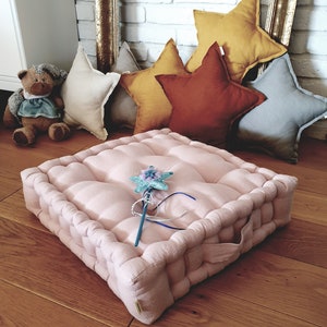 May include: A pink floor cushion with a star-shaped toy on top. The cushion is made of linen and has a tufted design. The cushion is surrounded by star-shaped pillows in various colors.