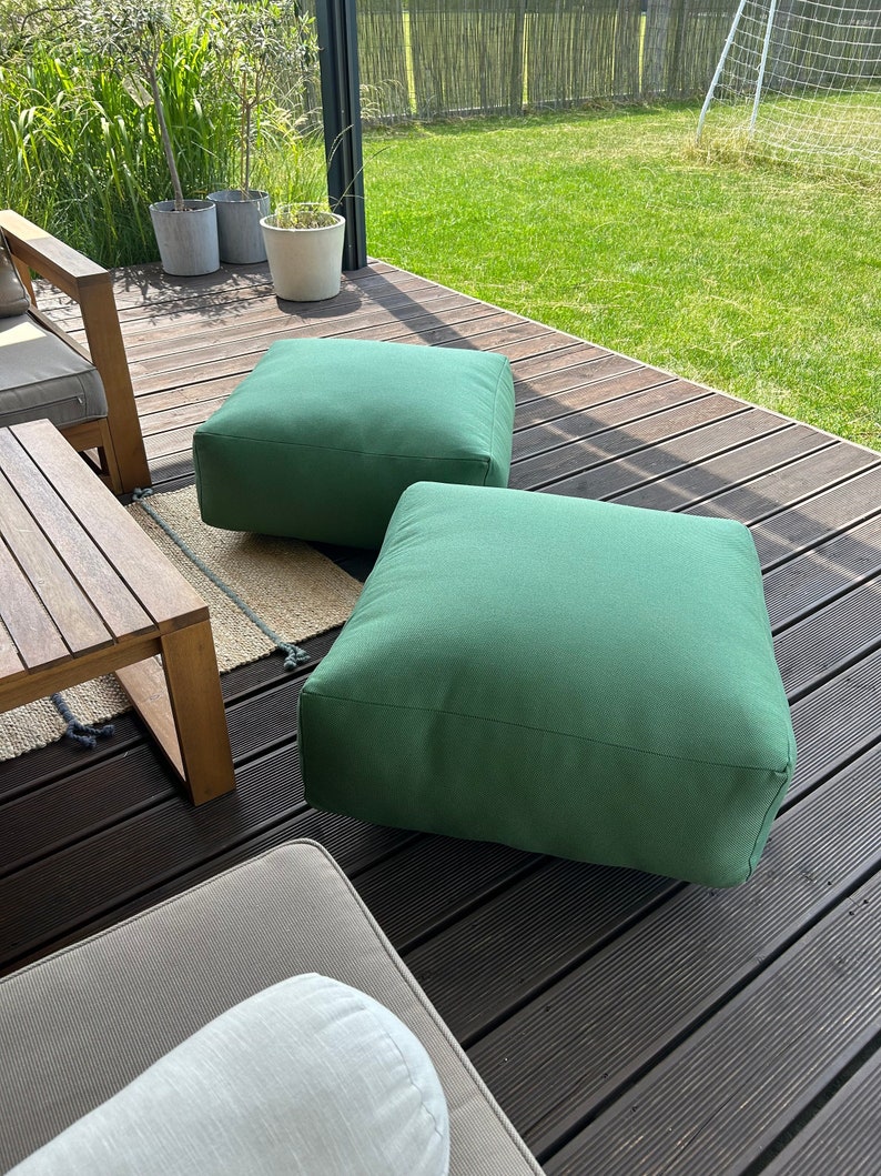 May include: Two green square floor cushions on a wooden deck. The cushions are made of a soft, textured fabric.