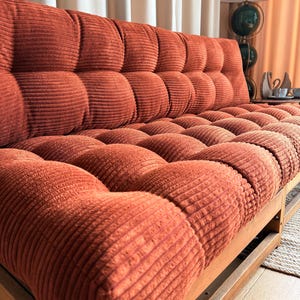 K&ouml;nnte beinhalten: Ein Sofa im Retro-Stil mit einem Cordstoff in einem verbrannten Orange. Das Sofa hat einen Holzrahmen und ein getuftetes Design auf der R&uuml;ckenlehne und den Sitzkissen. Das Sofa ist etwa 190 cm lang.