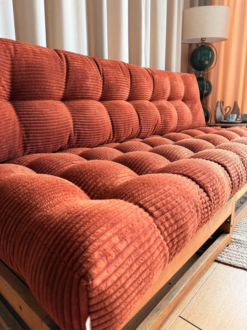 K&ouml;nnte beinhalten: Ein Sofa im Retro-Stil mit einem Cordstoff in einem verbrannten Orange. Das Sofa hat einen Holzrahmen und ein getuftetes Design auf der R&uuml;ckenlehne und den Sitzkissen. Das Sofa ist etwa 190 cm lang.
