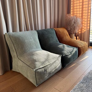 May include: Three corduroy lounge chairs in green and gold hues. The chairs have a low profile, rounded back, and square seat. They sit on a wooden floor, with a neutral curtain backdrop. The chairs are in shades of sage, teal, and mustard.