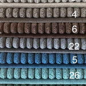 Op de afbeelding: Een stapel van 14 verschillende gekleurde stofstalen. De stalen zijn allemaal getextureerd en hebben een geribbeld patroon. De nummers 38, 100, 2, 3, 4, 6, 22, 5, 26, 14, 4, 46 en 44 zijn zichtbaar op de stalen.