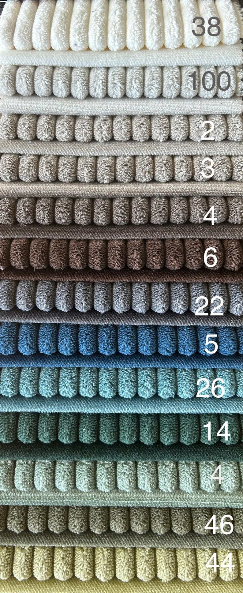 Op de afbeelding: Een stapel van 14 verschillende gekleurde stofstalen. De stalen zijn allemaal getextureerd en hebben een geribbeld patroon. De nummers 38, 100, 2, 3, 4, 6, 22, 5, 26, 14, 4, 46 en 44 zijn zichtbaar op de stalen.