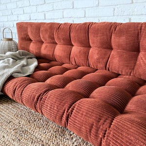 K&ouml;nnte beinhalten: Ein rostfarbenes Cord-Bodensofa mit getufteter R&uuml;ckenlehne und Sitzfl&auml;che. Eine hellgraue Decke ist &uuml;ber das Sofa drapiert. Das Sofa steht auf einem gewebten Jute-Teppich, mit einer wei&szlig;en Backsteinmauer im Hintergrund.