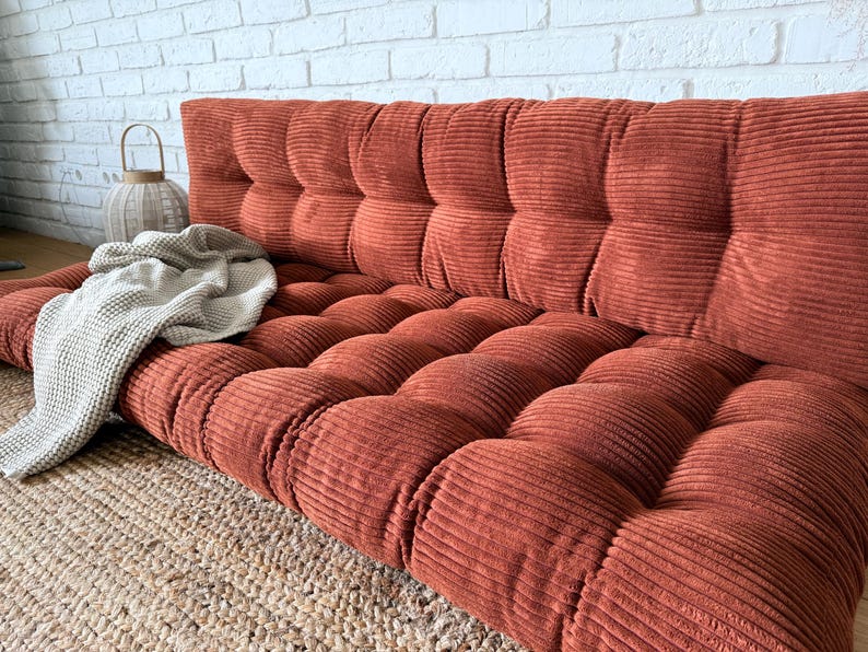 K&ouml;nnte beinhalten: Ein rostfarbenes Cord-Bodensofa mit getufteter R&uuml;ckenlehne und Sitzfl&auml;che. Eine hellgraue Decke ist &uuml;ber das Sofa drapiert. Das Sofa steht auf einem gewebten Jute-Teppich, mit einer wei&szlig;en Backsteinmauer im Hintergrund.