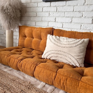 Op de afbeelding: Een mosterdgele corduroy bank met een wit gestoffeerd kussen en een wit kussen met franjes. De bank staat voor een witte bakstenen muur en een ingelijst zwart-witdruk.