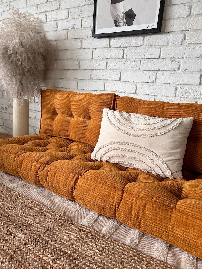 Op de afbeelding: Een mosterdgele corduroy bank met een wit gestoffeerd kussen en een wit kussen met franjes. De bank staat voor een witte bakstenen muur en een ingelijst zwart-witdruk.