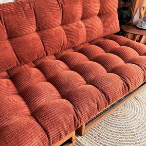 K&ouml;nnte beinhalten: Ein rostfarbenes Cord-Futon mit Holzrahmen. Das Futon hat ein getuftetes Design auf den R&uuml;cken- und Sitzkissen. Der Cordstoff hat eine gerippte Textur. Das Futon steht auf einem Holzboden neben einem runden, gewebten Teppich.