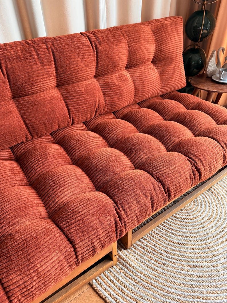K&ouml;nnte beinhalten: Ein rostfarbenes Cord-Futon mit Holzrahmen. Das Futon hat ein getuftetes Design auf den R&uuml;cken- und Sitzkissen. Der Cordstoff hat eine gerippte Textur. Das Futon steht auf einem Holzboden neben einem runden, gewebten Teppich.