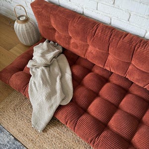 K&ouml;nnte beinhalten: Ein rostfarbenes Cordsofa mit einem gesteppten Design. Eine beige Strickdecke ist &uuml;ber dem Sofa drapiert. Ein gewebter Jute-Teppich und eine dekorative Laterne sind ebenfalls im Bild.