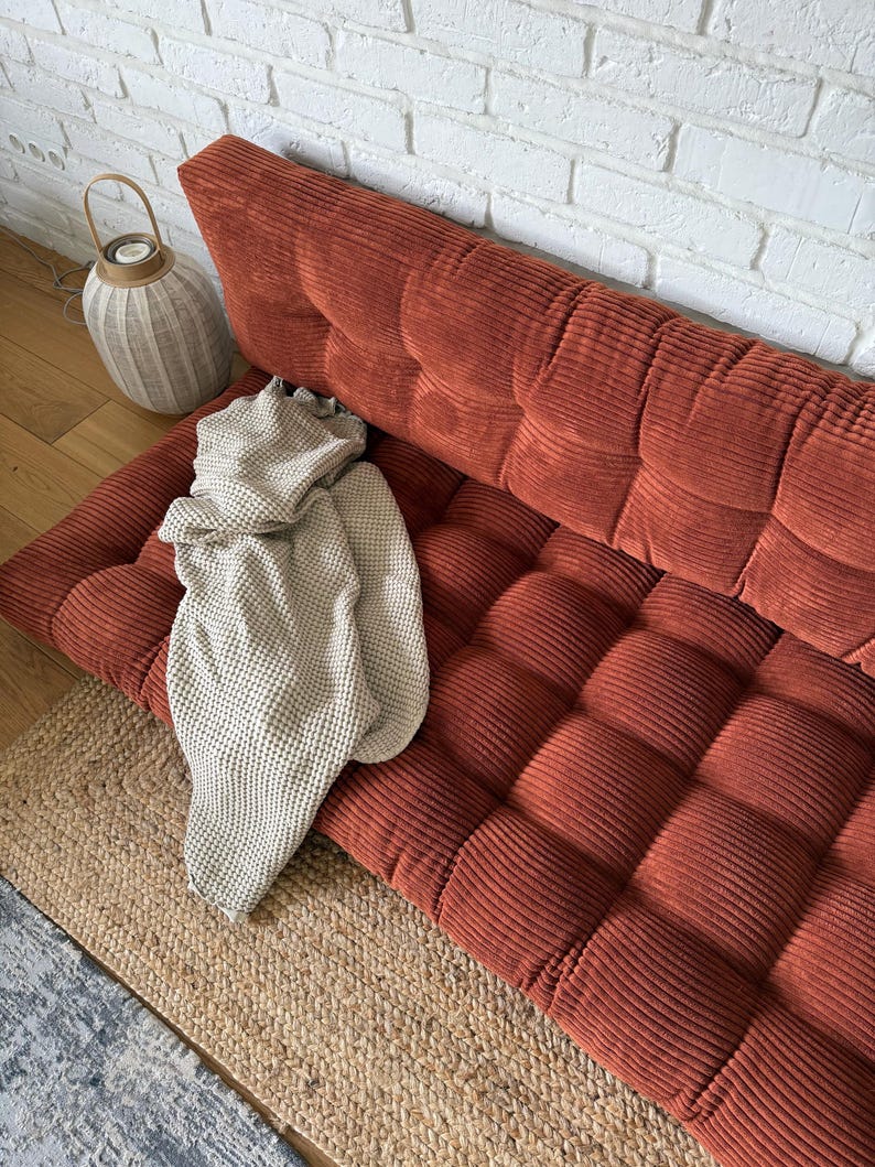 K&ouml;nnte beinhalten: Ein rostfarbenes Cordsofa mit einem gesteppten Design. Eine beige Strickdecke ist &uuml;ber dem Sofa drapiert. Ein gewebter Jute-Teppich und eine dekorative Laterne sind ebenfalls im Bild.