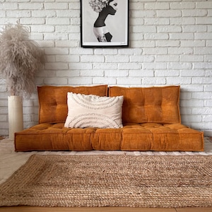 Op de afbeelding: Een mosterdgele corduroy vloercushion met een wit kussen en een zwart-wit ingelijste print van een vrouw met bloemen in haar haar, hangend aan een witte bakstenen muur.