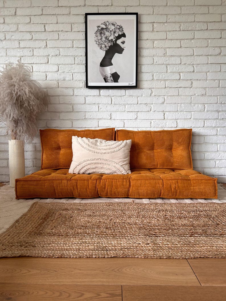 Op de afbeelding: Een mosterdgele corduroy vloercushion met een wit kussen en een zwart-wit ingelijste print van een vrouw met bloemen in haar haar, hangend aan een witte bakstenen muur.