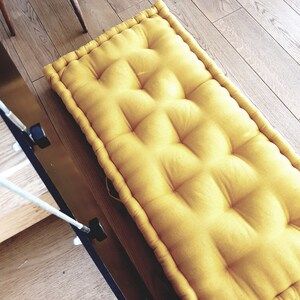 Op de afbeelding: Een gele vloercushion met een gestoffeerd ontwerp. De kussen is gemaakt van een zachte, comfortabele stof en heeft een eenvoudige, moderne stijl.