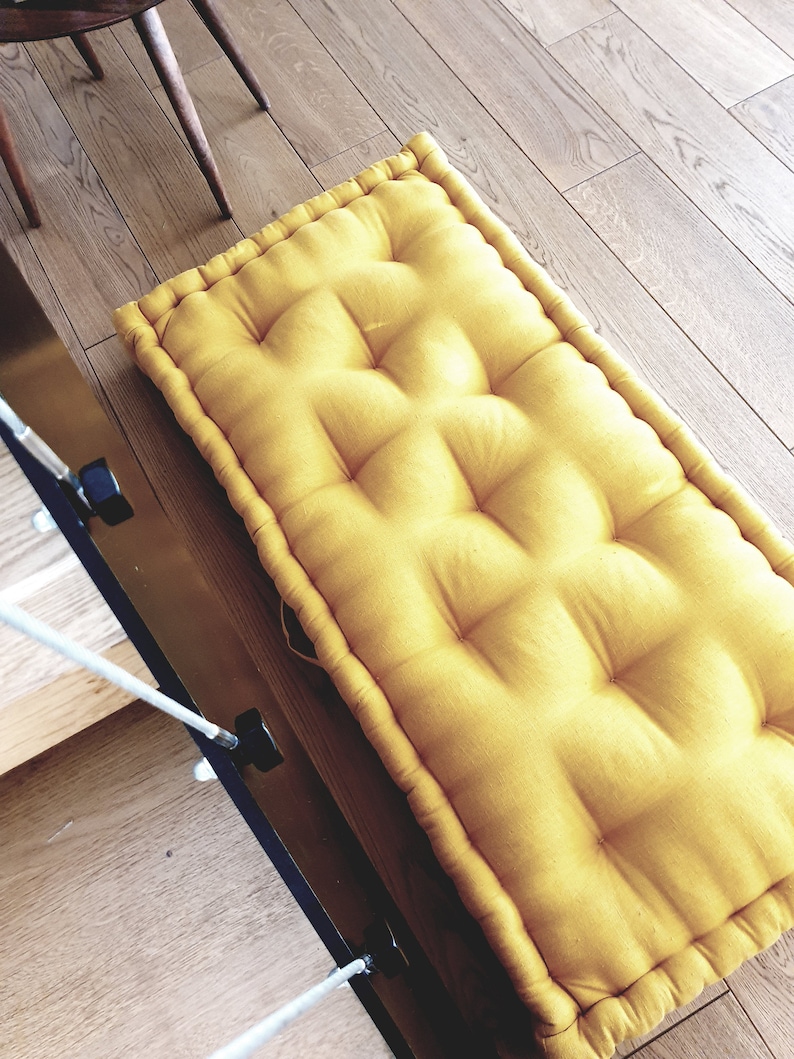 Op de afbeelding: Een gele vloercushion met een gestoffeerd ontwerp. De kussen is gemaakt van een zachte, comfortabele stof en heeft een eenvoudige, moderne stijl.