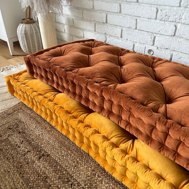 Peut inclure: Deux coussins de sol, l'un brun et l'autre jaune, avec un rembourrage capitonn&eacute;. Les coussins sont empil&eacute;s l'un sur l'autre et reposent sur un tapis tiss&eacute;.
