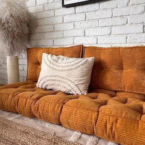 Puede incluir: Un cojín de suelo de pana de color mostaza con botones acolchados y una almohada blanca con ribete de flecos. El cojín está sobre una alfombra marrón claro frente a una pared de ladrillo blanco.