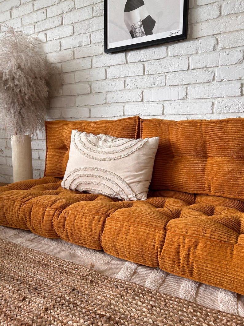 Op de afbeelding: Een mosterdgele corduroy vloercushion met knopen en een wit kussen met franjes. De cushion ligt op een lichtbruin tapijt voor een witte bakstenen muur.