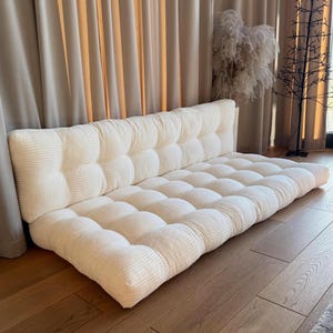 May include: A cream-colored, tufted floor sofa with a corduroy texture. The sofa has a rectangular shape with a backrest and a cushioned seat. The neutral color and simple design make it suitable for various interior styles.