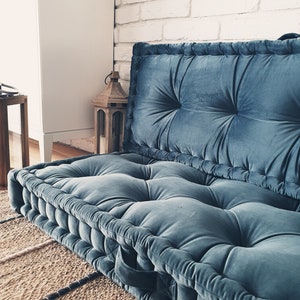 May include: A large, blue, tufted floor cushion with a white brick wall in the background.