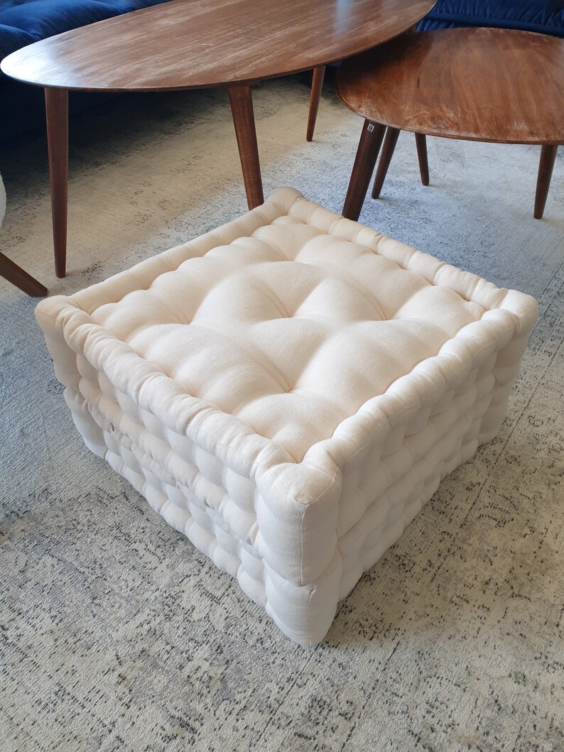 May include: A white, tufted, square floor cushion with a linen-like fabric. The cushion is made of multiple layers and has a thick, comfortable appearance.