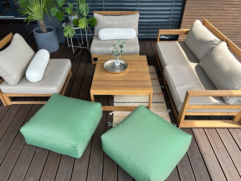 May include: A wooden outdoor furniture set with a sofa, two chairs, a coffee table, and two green ottomans. The furniture is arranged on a wooden deck with a rug in the centre.