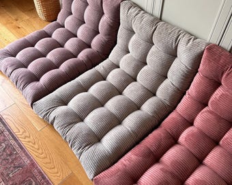 May include: Three floor cushions in different colors: purple, gray, and pink. The cushions are made of a soft, textured fabric and have a tufted design.