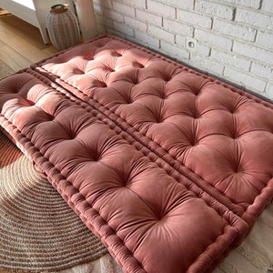 Peut inclure: Deux coussins de sol en velours rose avec des boutons capitonnés et un bord épais piqué. Les coussins sont disposés en quinconce sur un sol en bois brun clair.