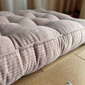 May include: A close-up of a light pink corduroy cushion with a tufted design. The cushion is sitting on a wooden surface.