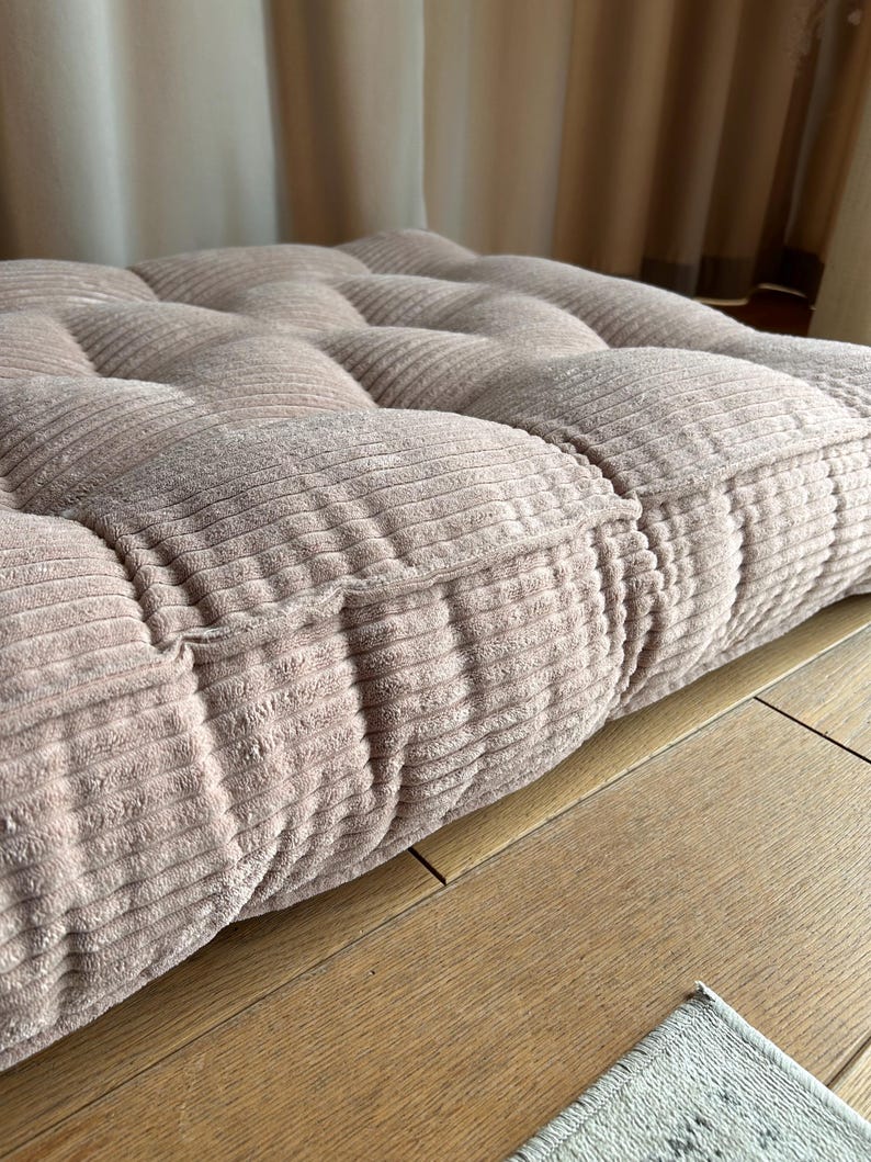 May include: A close-up of a light pink corduroy cushion with a tufted design. The cushion is sitting on a wooden surface.
