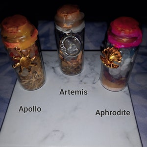 May include: Three small glass bottles with cork tops and various charms and crystals inside. The bottles are labelled "Apollo", "Artemis", and "Aphrodite".
