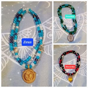 Könnte beinhalten: Drei Perlenketten mit Anhängern. Die erste Kette ist blau und weiß mit einem goldenen Anhänger, auf dem "Zeus" steht. Die zweite Kette ist grün und weiß mit einem goldenen Dreizack-Anhänger, auf dem "Poseidon" steht. Die dritte Kette ist rot und schwarz mit einem goldenen Anhänger, auf dem "Discordia" steht.