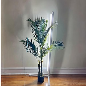 May include: A white floor lamp with a long, thin, white light source. The lamp is next to a potted green palm tree.