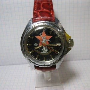 Puede incluir: Un reloj de pulsera negro y plateado con una estrella roja y una mujer sosteniendo una espada en la esfera. El reloj tiene una correa de cuero roja y el texto "1995" en la esfera.