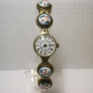 Puede incluir: Reloj de pulsera vintage dorado con esfera blanca y números romanos. La correa del reloj presenta eslabones ovalados decorativos con diseños florales en rosa, blanco y azul. La esfera del reloj tiene el texto "ЧАЙКА" y "17 jewels".