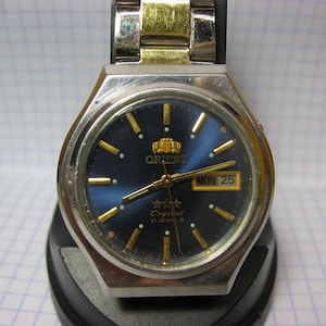 May include: A silver and gold Orient watch with a blue face and gold hands. The watch face has a date window that reads "MON 25" and the text "Crystal 21 Jewels".