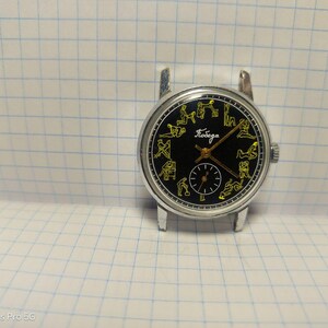 May include: A vintage wristwatch with a black face and silver casing. The watch face features yellow figures around the perimeter and a smaller subdial. The word "Победа" is written in Cyrillic script. The watch is set against a grid background.