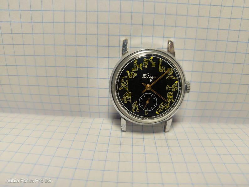 May include: A vintage wristwatch with a black face and silver casing. The watch face features yellow figures around the perimeter and a smaller subdial. The word "Победа" is written in Cyrillic script. The watch is set against a grid background.