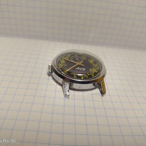 May include: A vintage wristwatch with a black face and gold hands and accents. The watch has a silver-colored case and a clear crystal. The watch is resting on a white grid-lined surface.