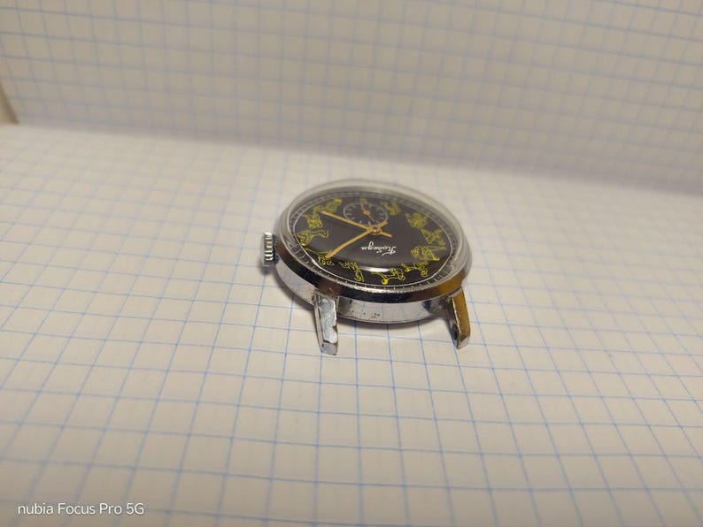 May include: A vintage wristwatch with a black face and gold hands and accents. The watch has a silver-colored case and a clear crystal. The watch is resting on a white grid-lined surface.