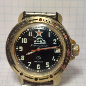 Puede incluir: Un reloj de pulsera vintage con esfera negra, bisel dorado y un emblema de estrella roja. El reloj presenta números blancos, una ventana de fecha y el texto "Каисндурские". El bisel está adornado con detalles rojos.