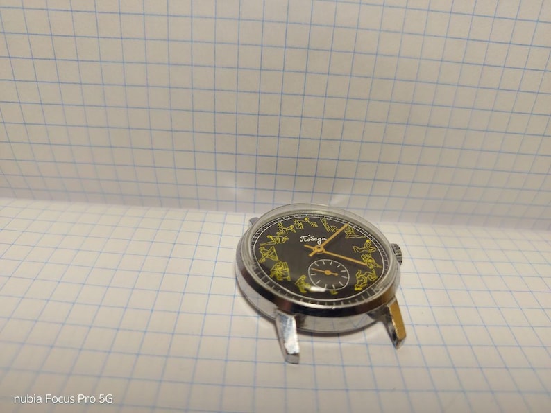 May include: A vintage wristwatch with a black face and gold hands and markers. The watch has a silver-colored metal case and a small subdial. The face features the word "Победа" in gold lettering.